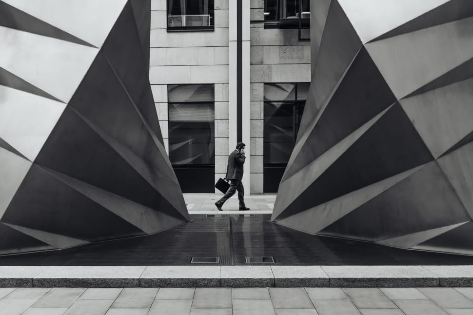 grayscale photography of man in formal attire holding handbag and using phone while walking beside concrete structure during daytime preview