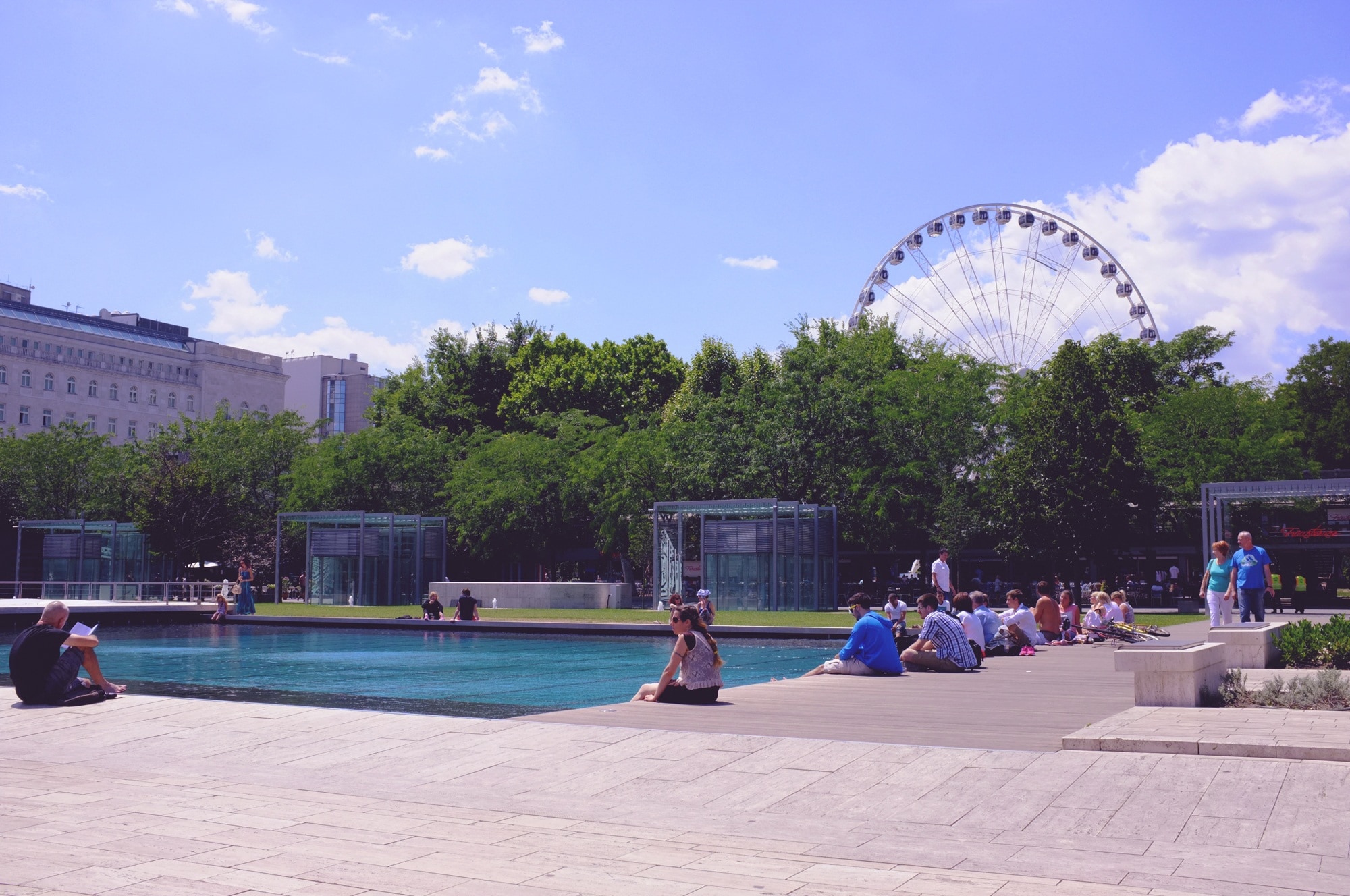 blue swimming pool; beige concrete park; green trees; beige concrete building; ferris wheel