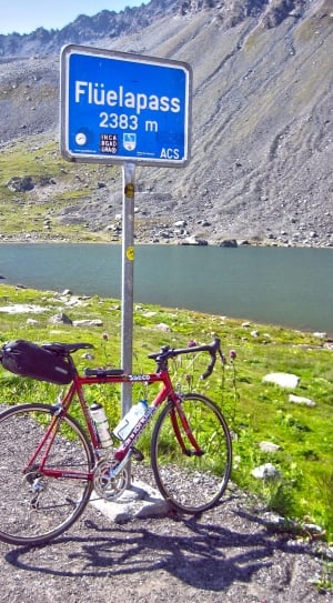 black and red road bike leaning on grey and blue Fluelapass signage near body of water thumbnail