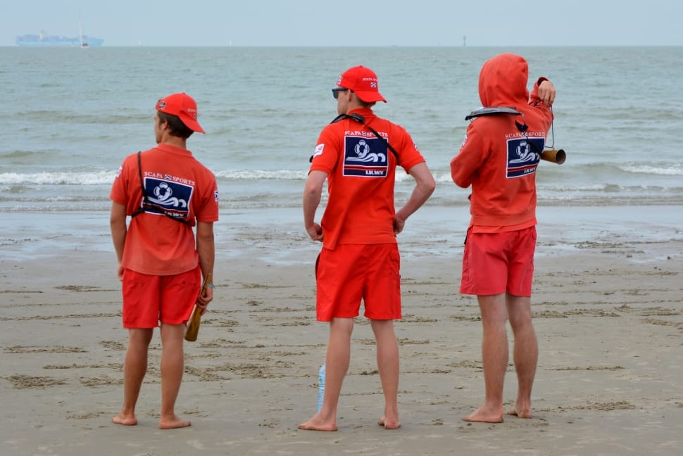 three men in red polo shirt and red shorts standing at the seashore during daytime preview