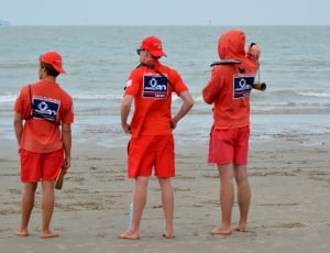 three men in red polo shirt and red shorts standing at the seashore during daytime thumbnail