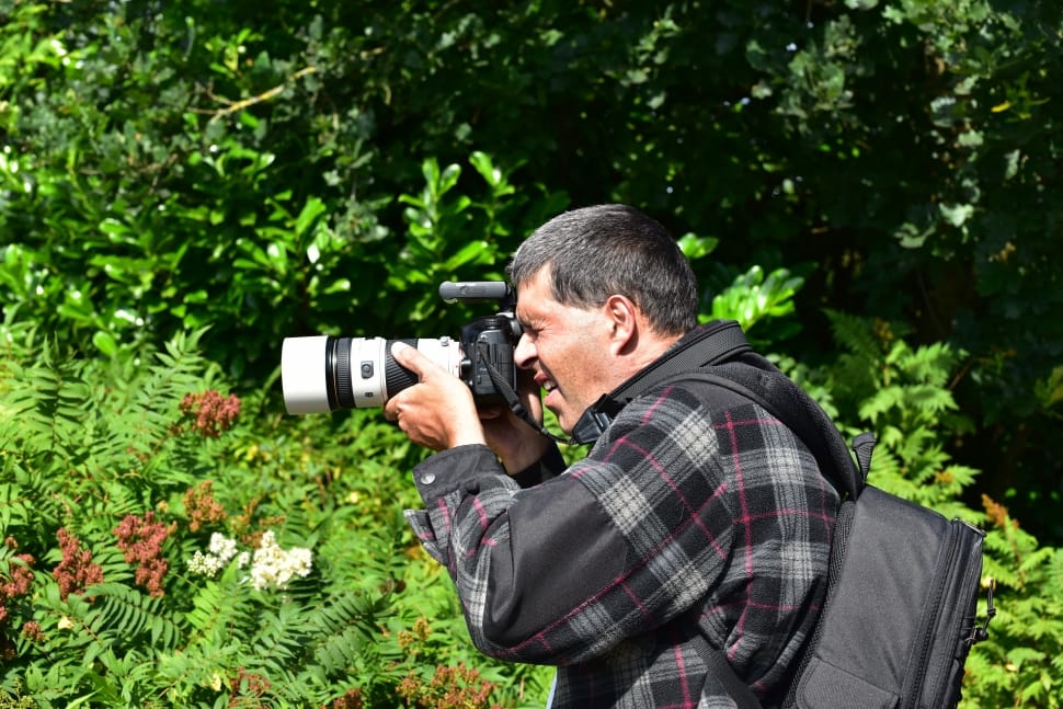 man in grey-and-pink plaid jacket with backpack holding white-and-black DSLR camera on hand overlooking plants during daytime preview
