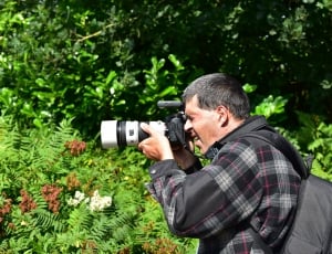 man in grey-and-pink plaid jacket with backpack holding white-and-black DSLR camera on hand overlooking plants during daytime thumbnail
