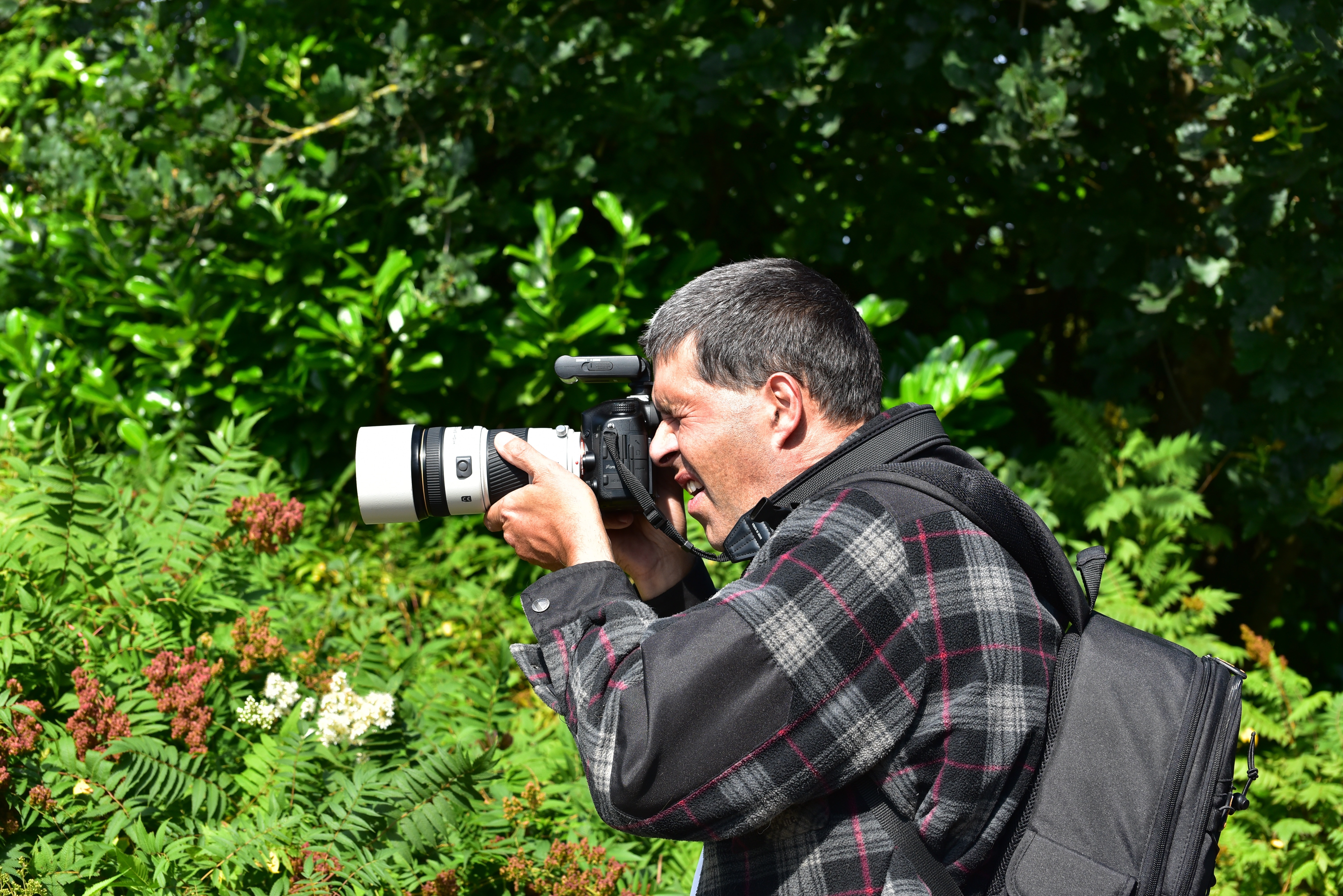 man in grey-and-pink plaid jacket with backpack holding white-and-black DSLR camera on hand overlooking plants during daytime