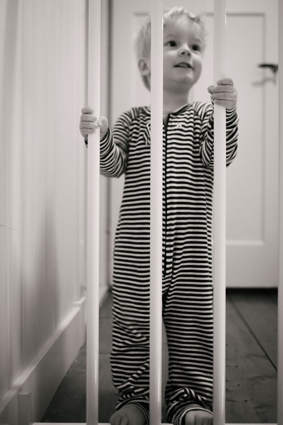 grayscale photo of toddler wearing stripe onesie sleeper holding white safety gate preview
