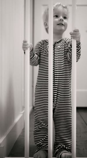 grayscale photo of toddler wearing stripe onesie sleeper holding white safety gate thumbnail