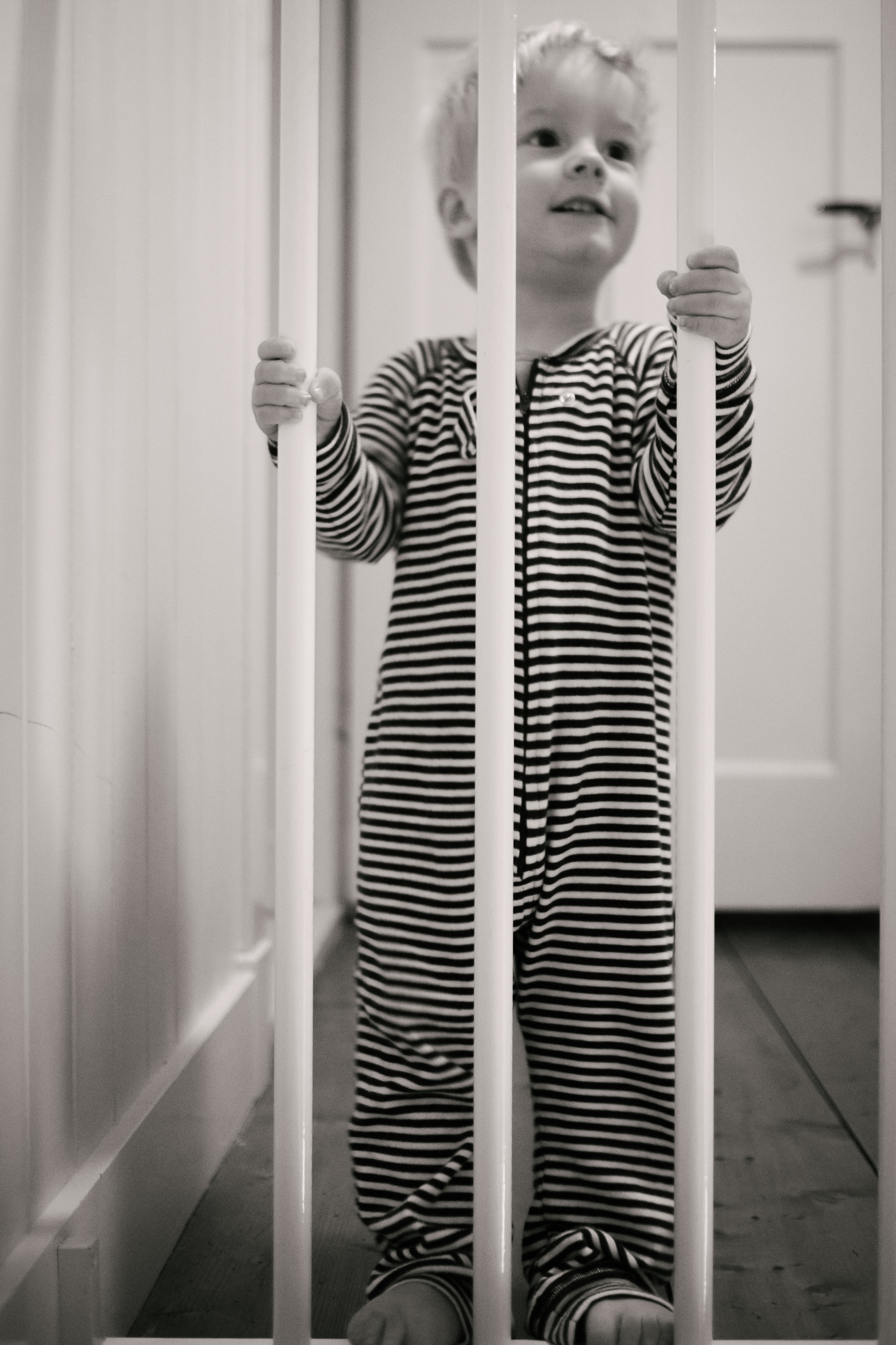 grayscale photo of toddler wearing stripe onesie sleeper holding white safety gate
