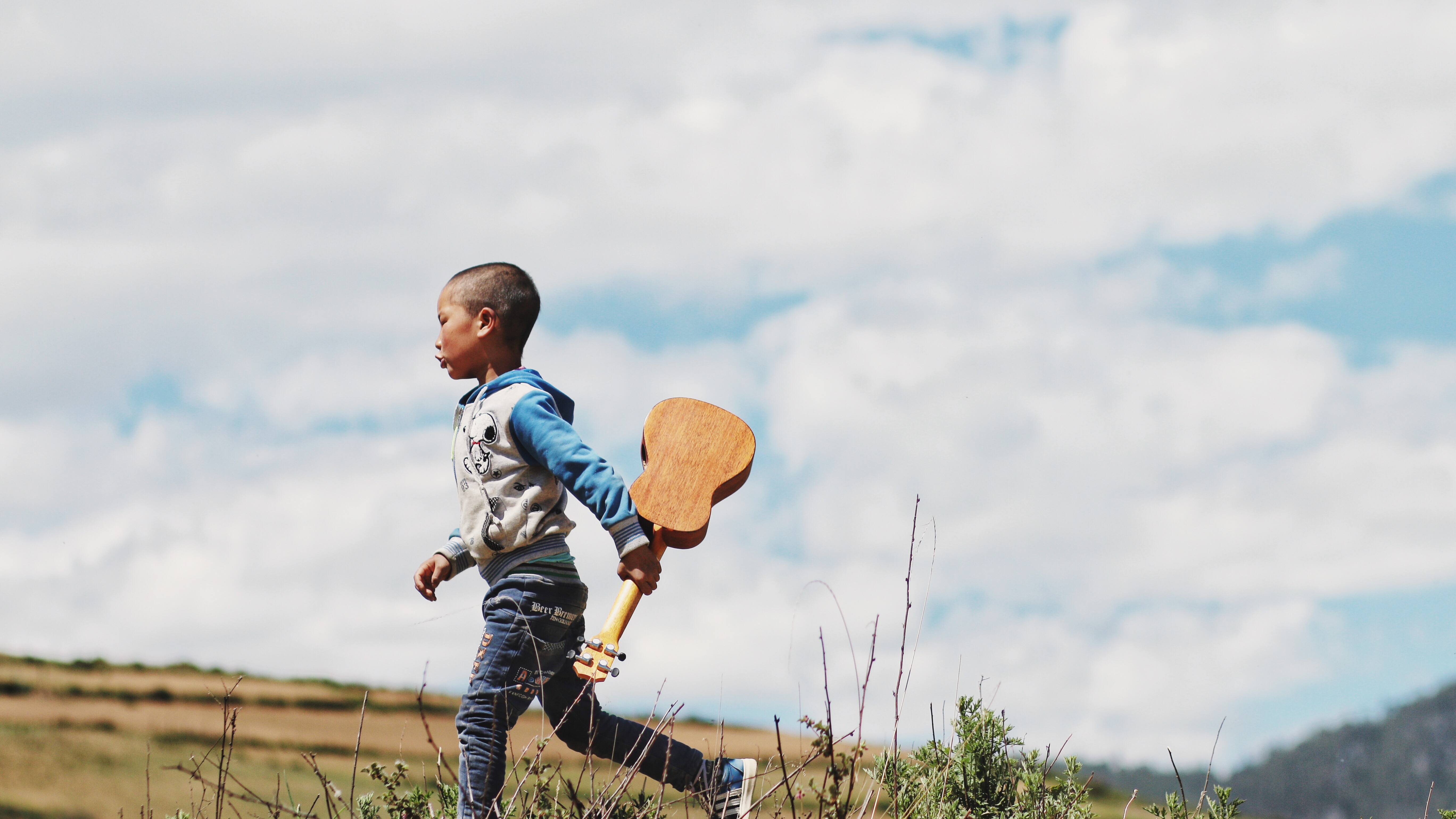 toddler wearing white and blue jacket holding brown ukulele walking on grass