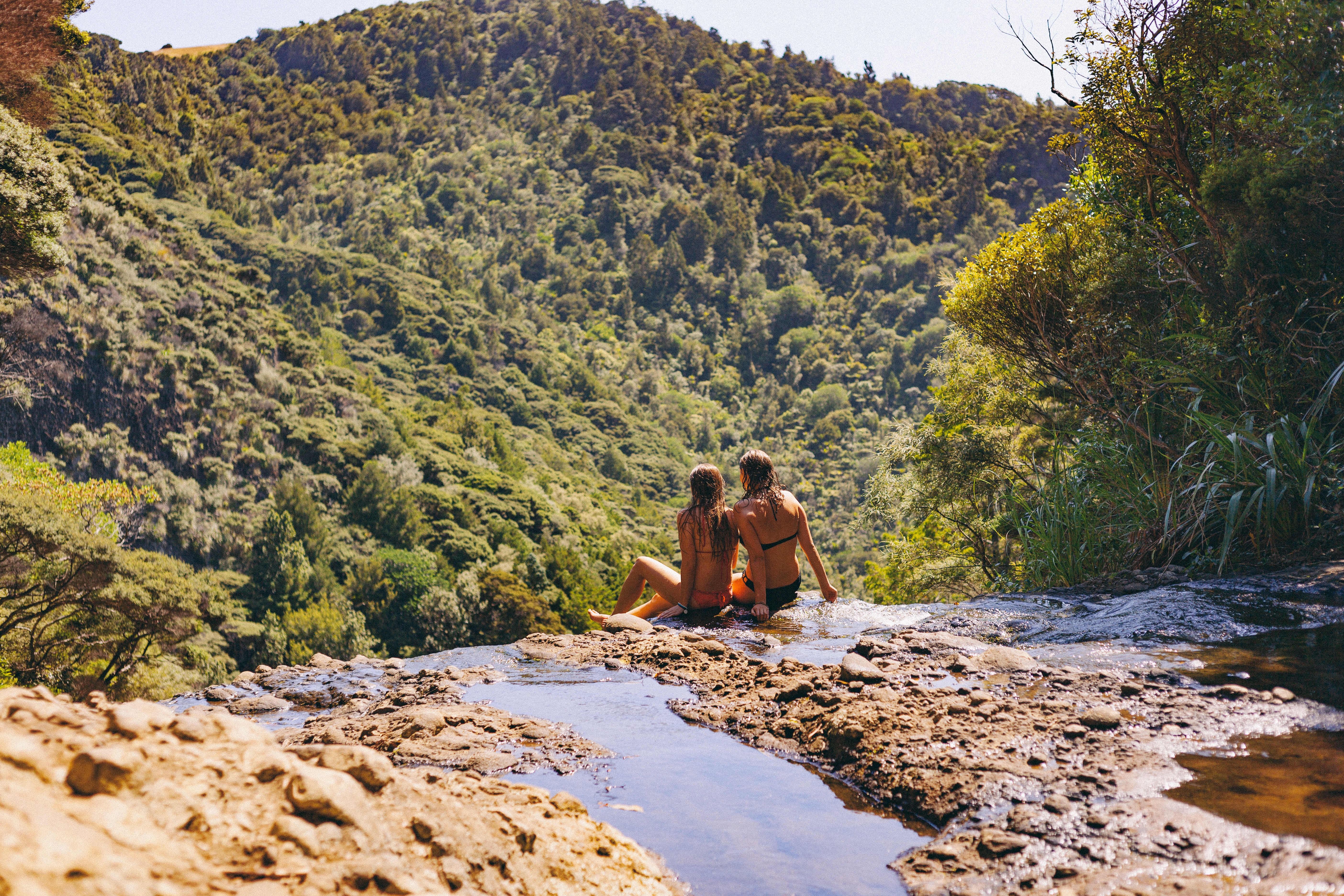 2 women wearing two piece bikini sitting on cliff at daytime