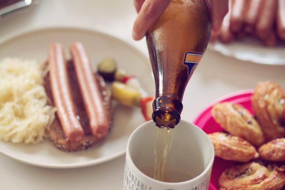 person filling white mug with beer next to sausage dish and brown pastries in plate preview