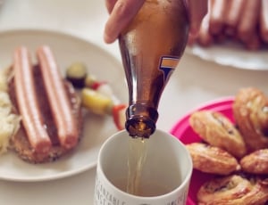 person filling white mug with beer next to sausage dish and brown pastries in plate thumbnail