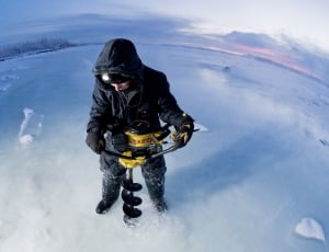 man wearing black jacket while using ice auger to pierce ice floor thumbnail