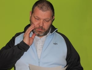 man in teal and black zip up jacket with hand on right cheek looking down at paper in other hand thumbnail