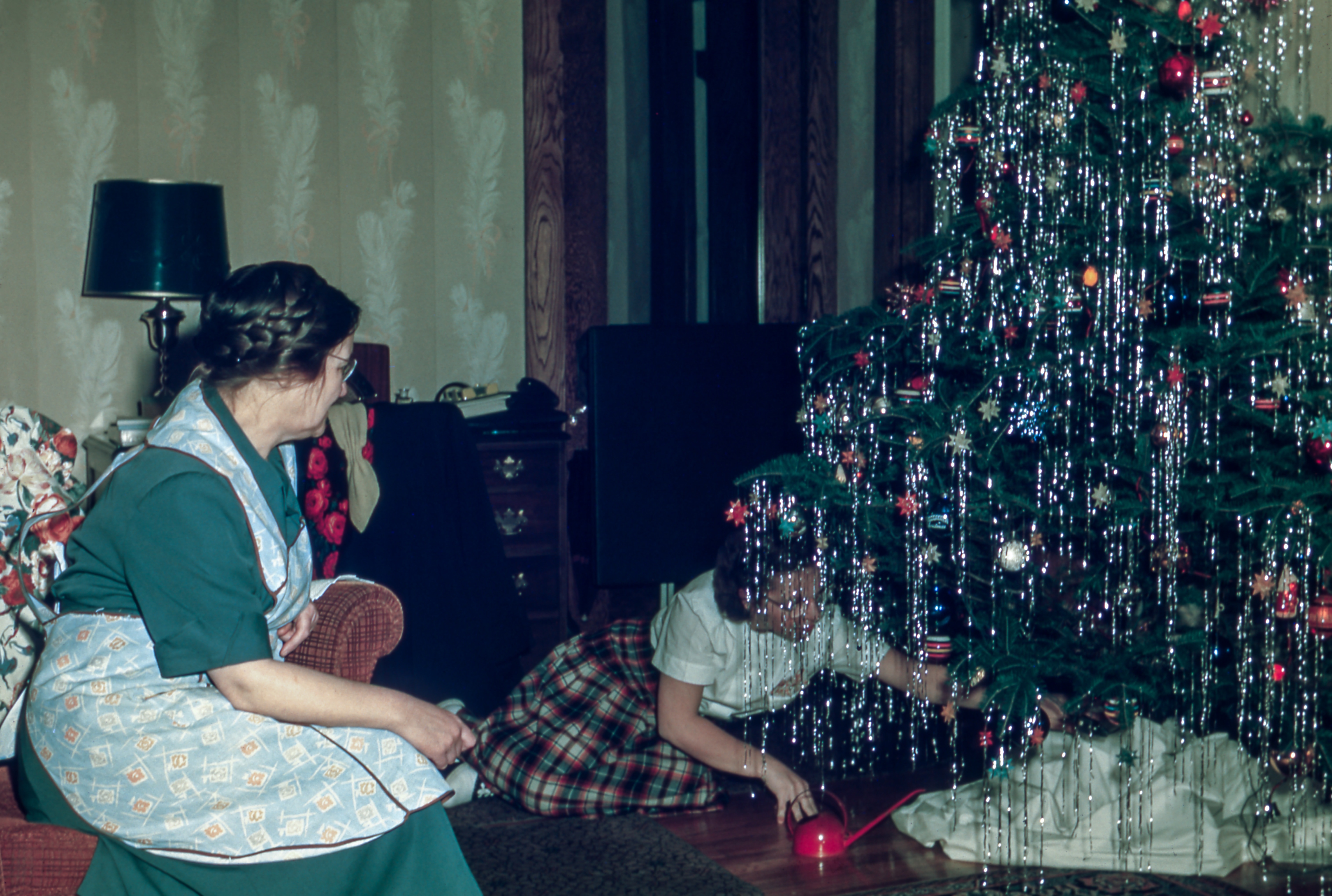 girl decorating the christmas tree while the woman sitting on a chair watching her