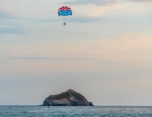 brown stone island in center of ocean red and blue parachute in sky thumbnail
