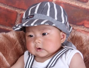 baby boy in white tank top and gray hat sitting on chair thumbnail