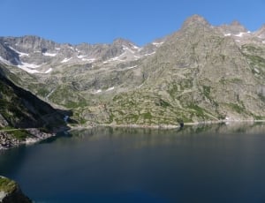 calm body of water surrounded by gray rocky mountain under clear blue sky during daytime thumbnail