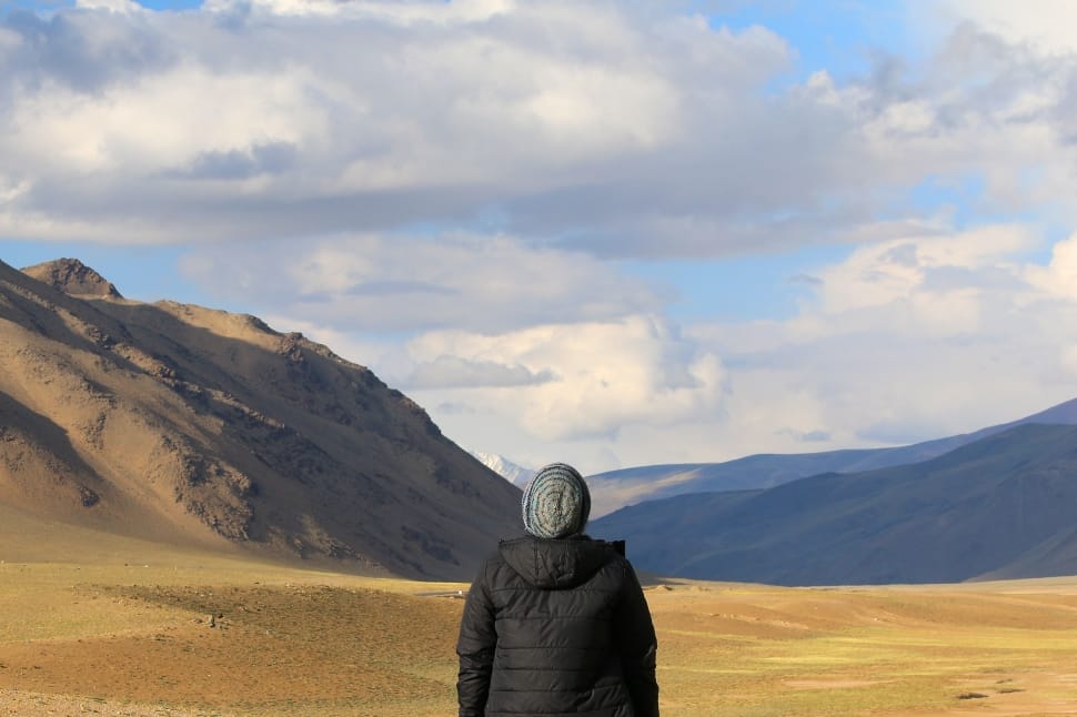person in black bubble hoodie wearing grey knit cap standing on brown dirt road near rock formation during daytime preview