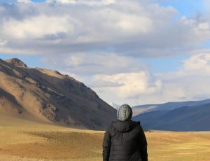 person in black bubble hoodie wearing grey knit cap standing on brown dirt road near rock formation during daytime thumbnail