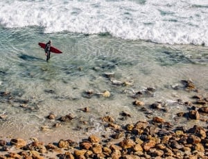 Person Holding Red Surfing Board in Clear Water Near Brown Stone during Daytime thumbnail