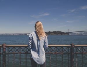 blonde haired woman in blue denim top standing near body of water during daytime thumbnail