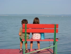 girl and boy on red bench near sea during daytime thumbnail