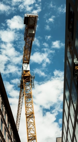 low angle photo of yellow crane in between two high rise buildings under blue sky and white clouds thumbnail