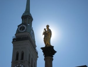 woman carrying baby statue near tower with analog clock under blue sky thumbnail