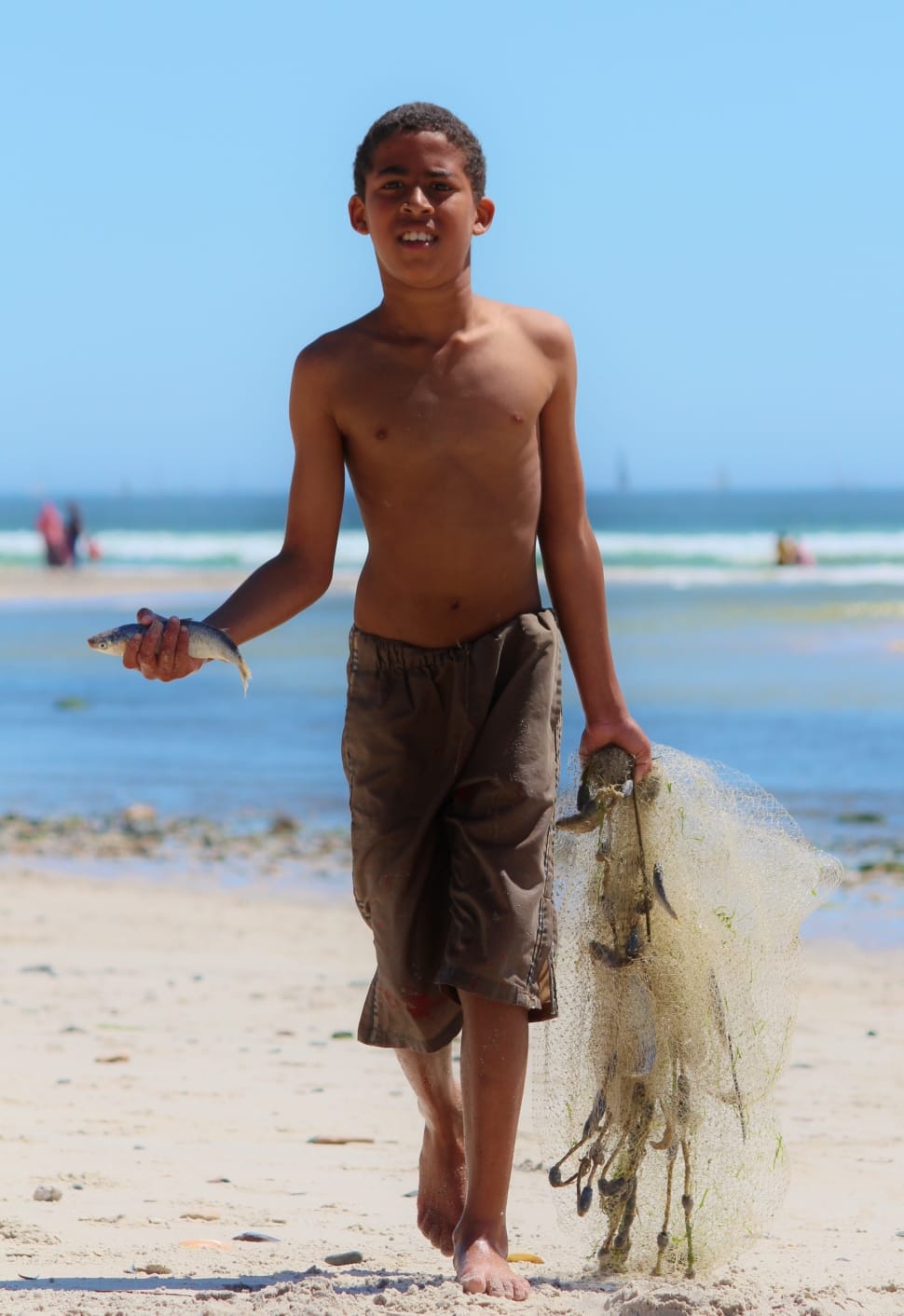 topless boy wearing gray shorts holding fish and net walking on sand during daytime preview