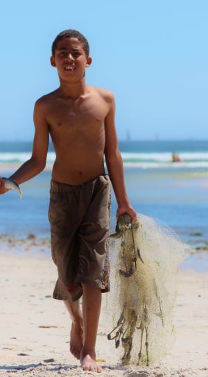 topless boy wearing gray shorts holding fish and net walking on sand during daytime thumbnail
