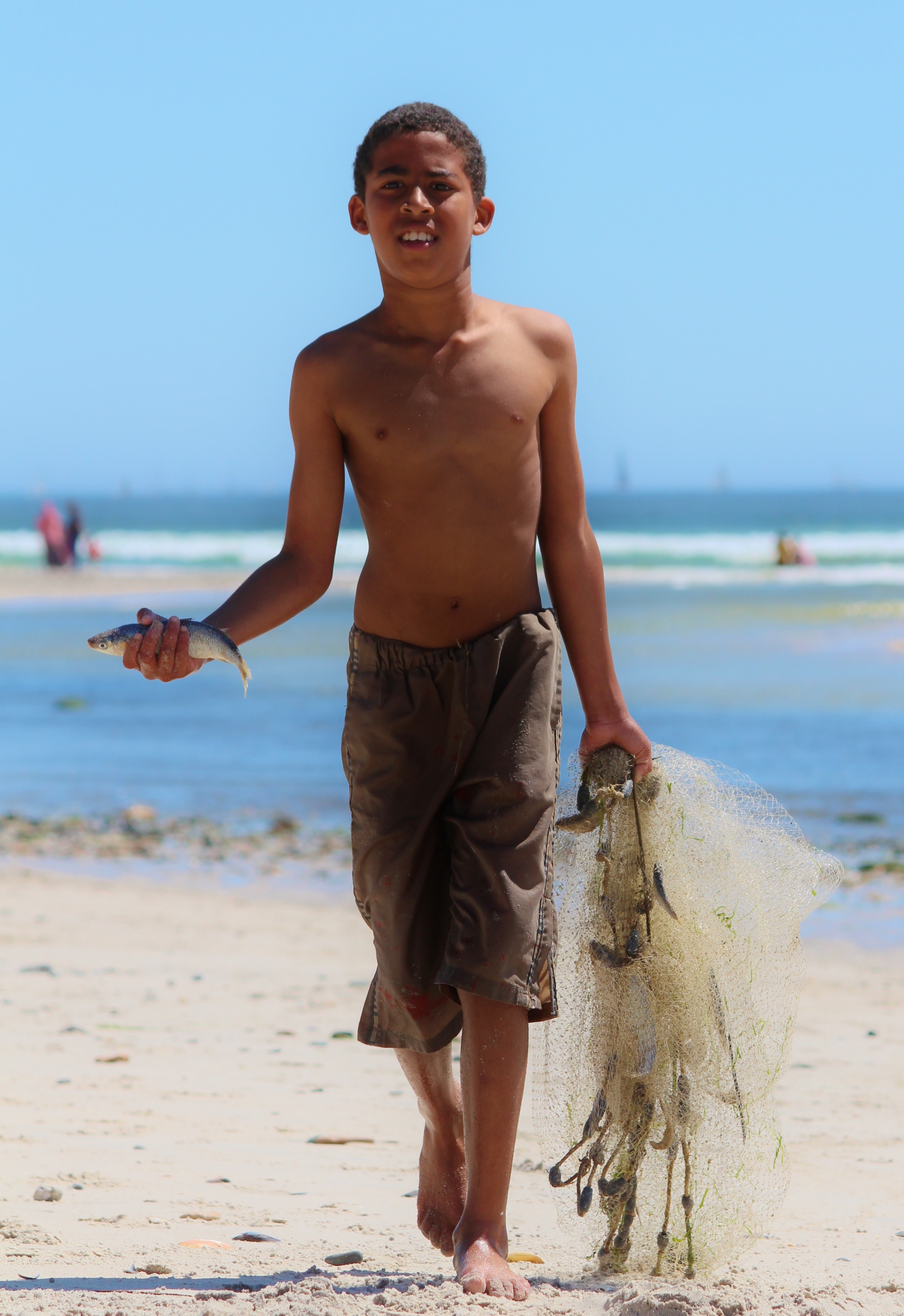 topless boy wearing gray shorts holding fish and net walking on sand during daytime
