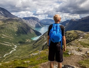 man in black shirt and short carrying blue backpack standing on mountain summit during day time thumbnail