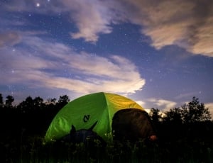 green and yellow tent surrounded with trees under a cloudy and starry sky during nighttime thumbnail
