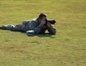man in camouflage soldier uniform holding bridge camera lying on grass at daytime thumbnail