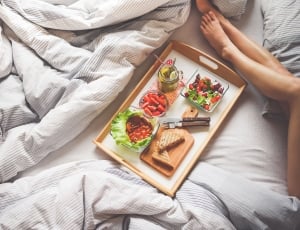 person laying in bed near brown serving tray filled with fruits and sandwich thumbnail