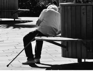 greyscale image of man with walking cane by his side sitting on bench under the sun thumbnail