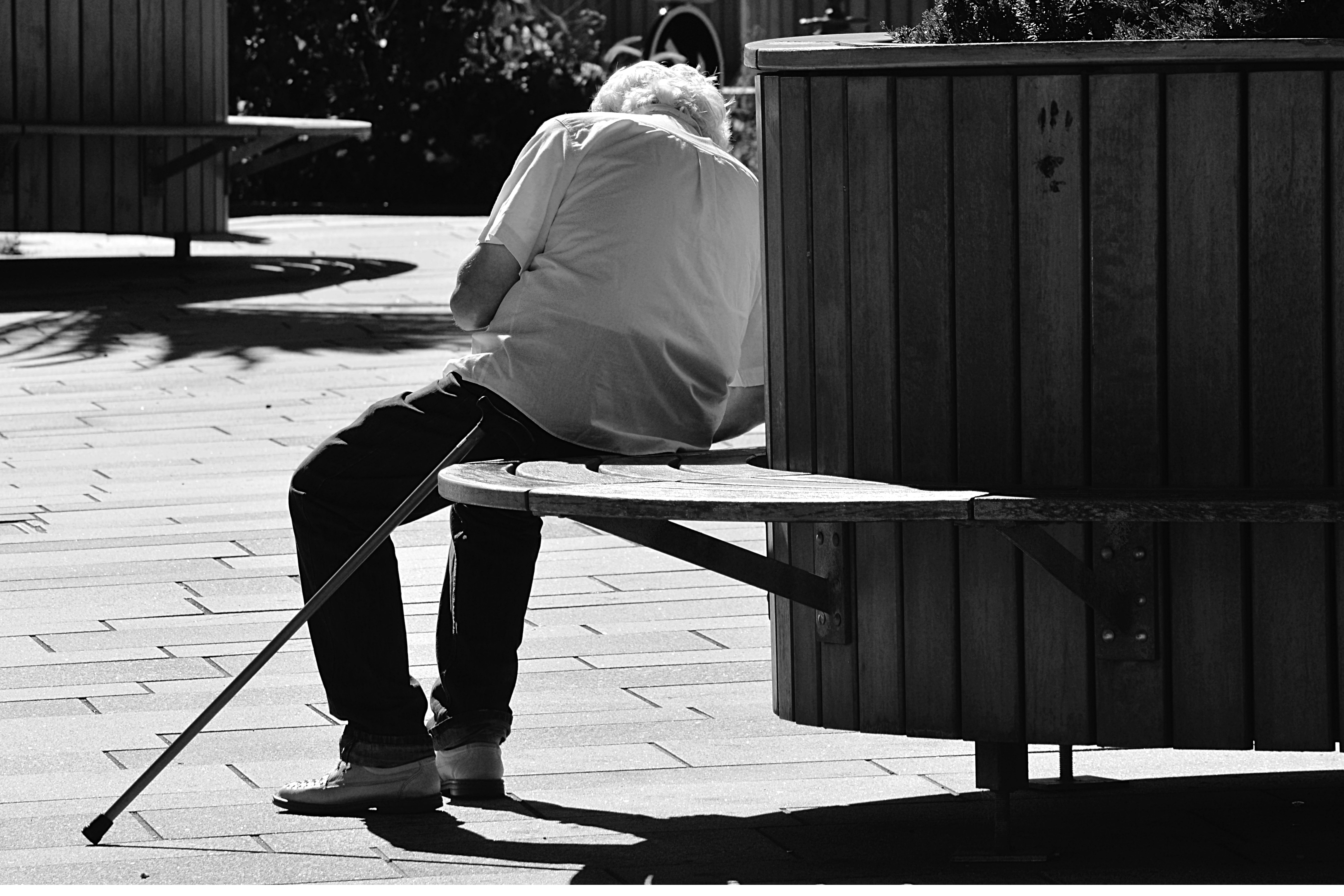 greyscale image of man with walking cane by his side sitting on bench under the sun