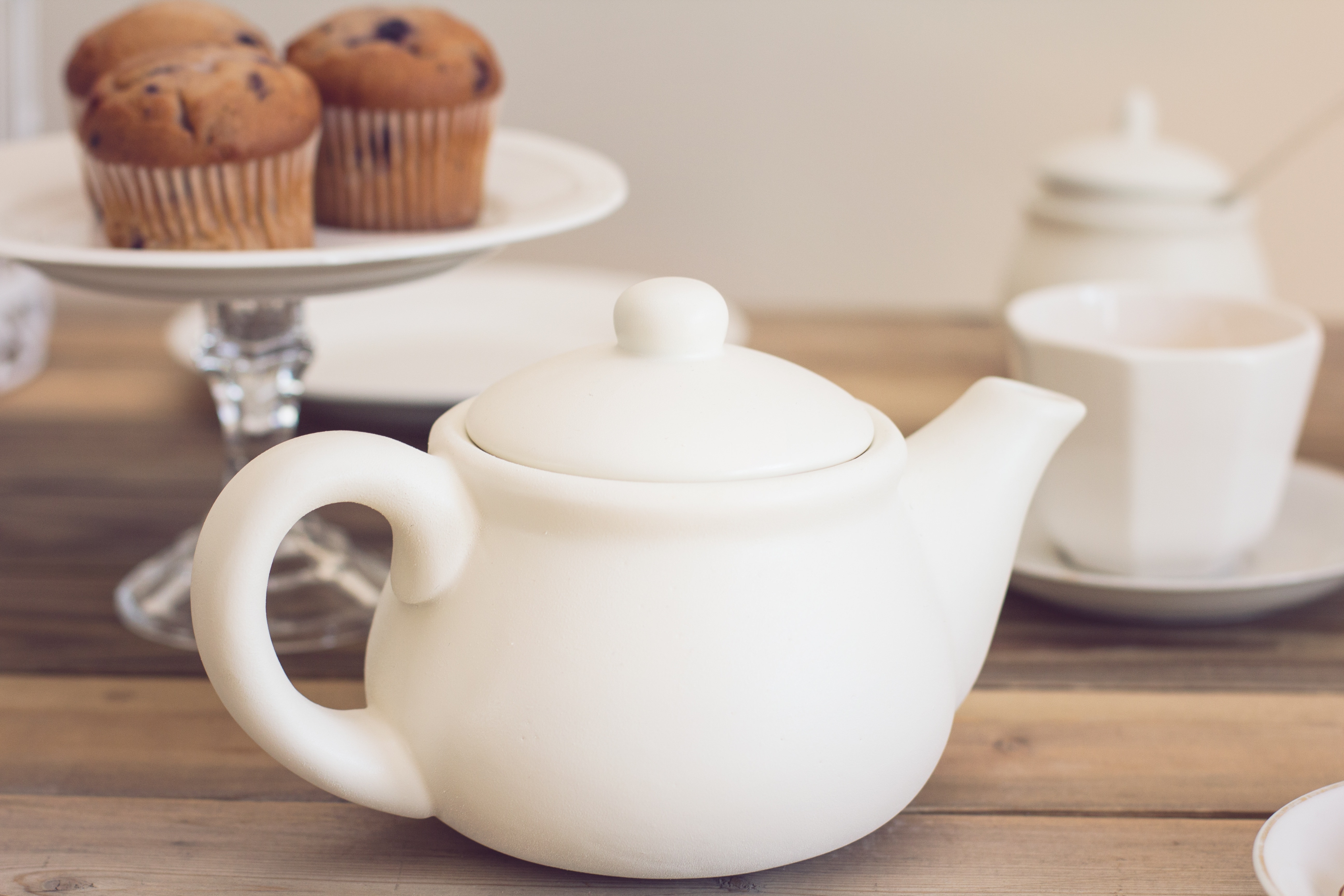 selective focus photo of white ceramic tea pot near three brown muffins on white plate