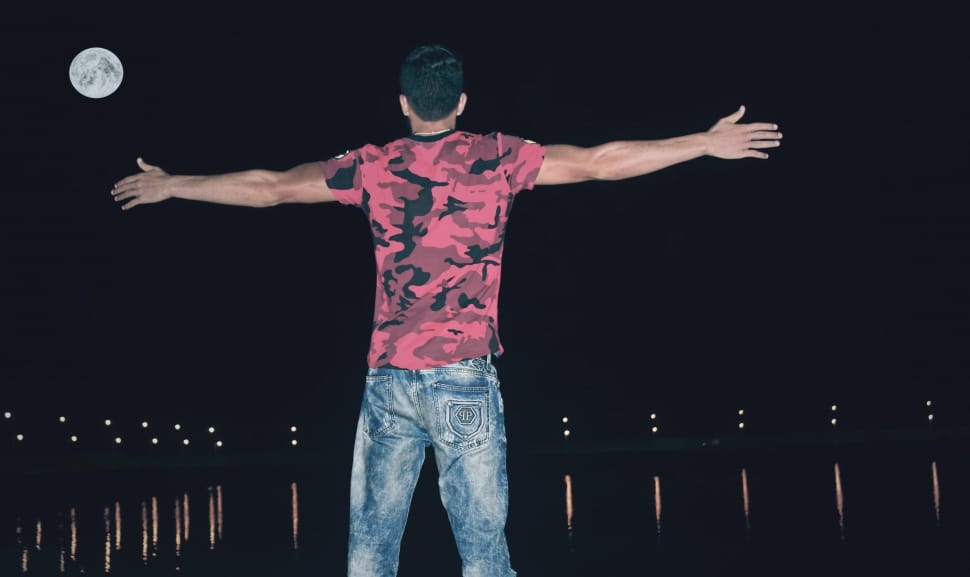 man in red and black camouflage shirt and jeans open arms near to body of water during night time preview