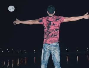 man in red and black camouflage shirt and jeans open arms near to body of water during night time thumbnail
