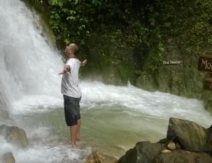 man wearing white shirt and black shorts standing near waterfalls thumbnail