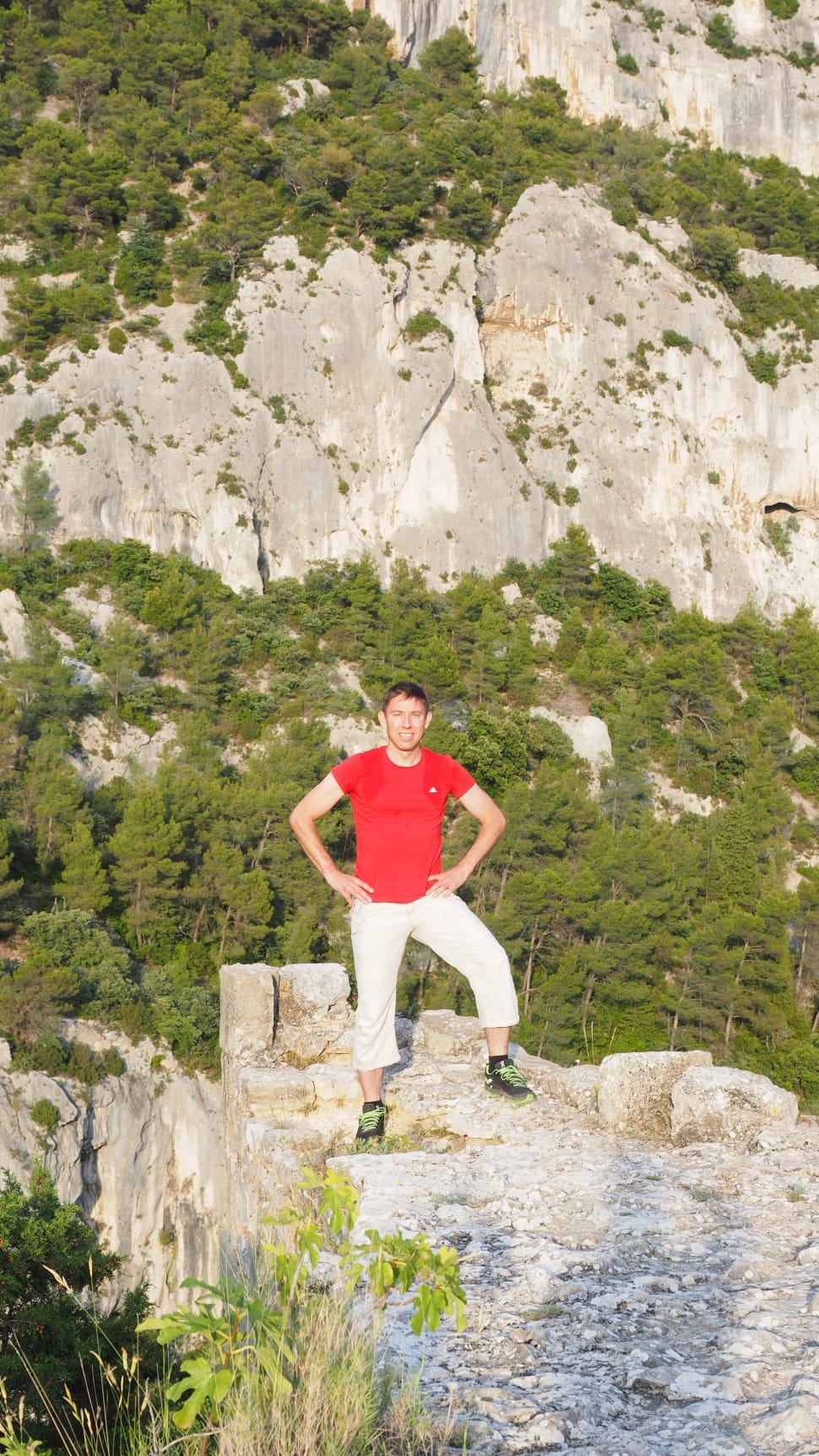 man in red shirt and white pants standing on cliff rock preview